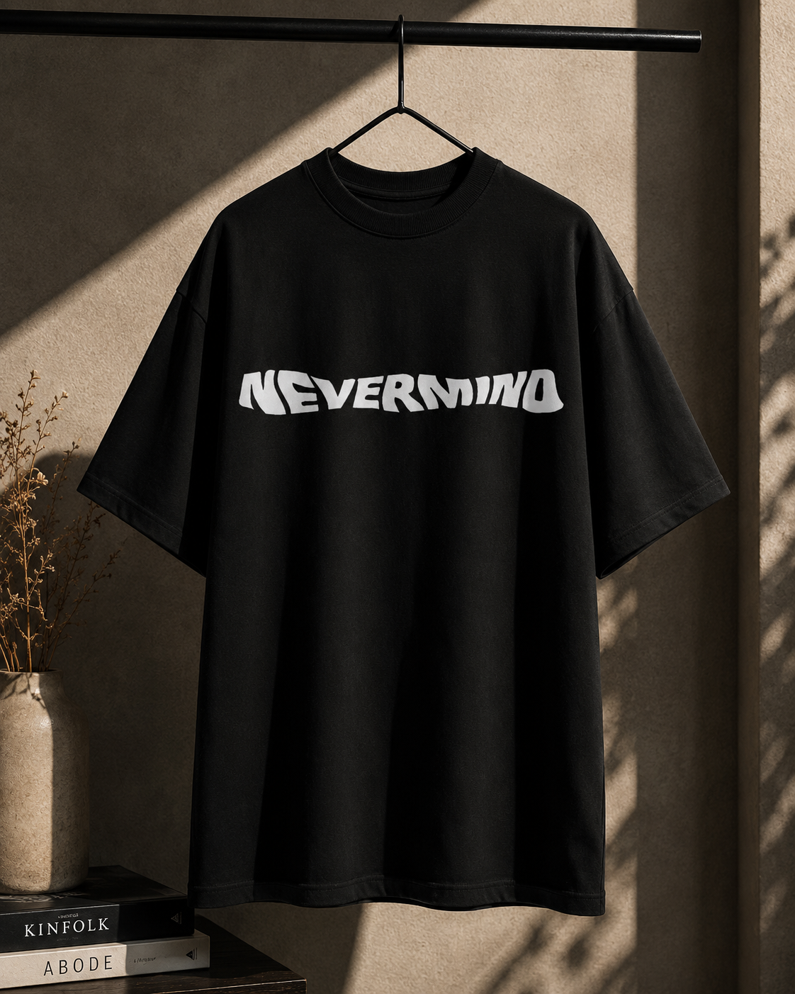 A black oversized short-sleeve t-shirt hanging on a black hanger from a clothing rack, featuring a stylized white 'NEVERMIND' graphic across the chest. The shirt is styled in a warm, sunlit room with a textured beige wall, a vase with dried stems, and books on a table.
