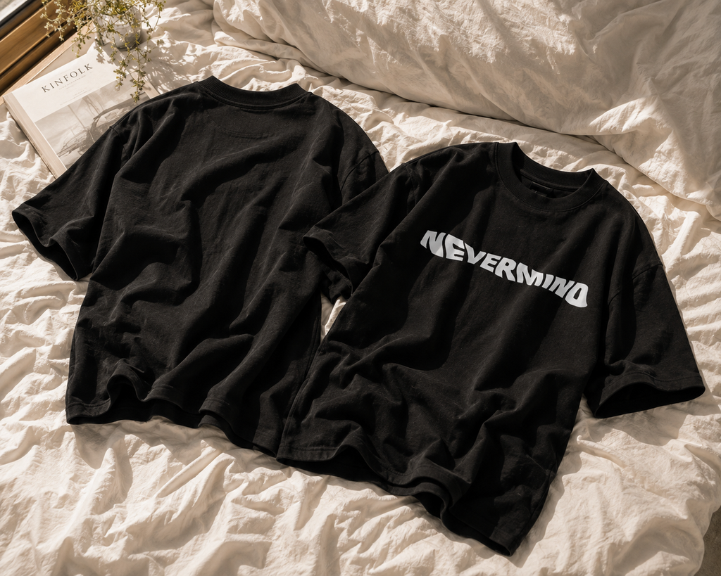 Two black oversized T-shirts laid flat on rumpled white bedding, showing the front and back. The front shirt has a large white 'NEVERMIND' graphic across the chest.