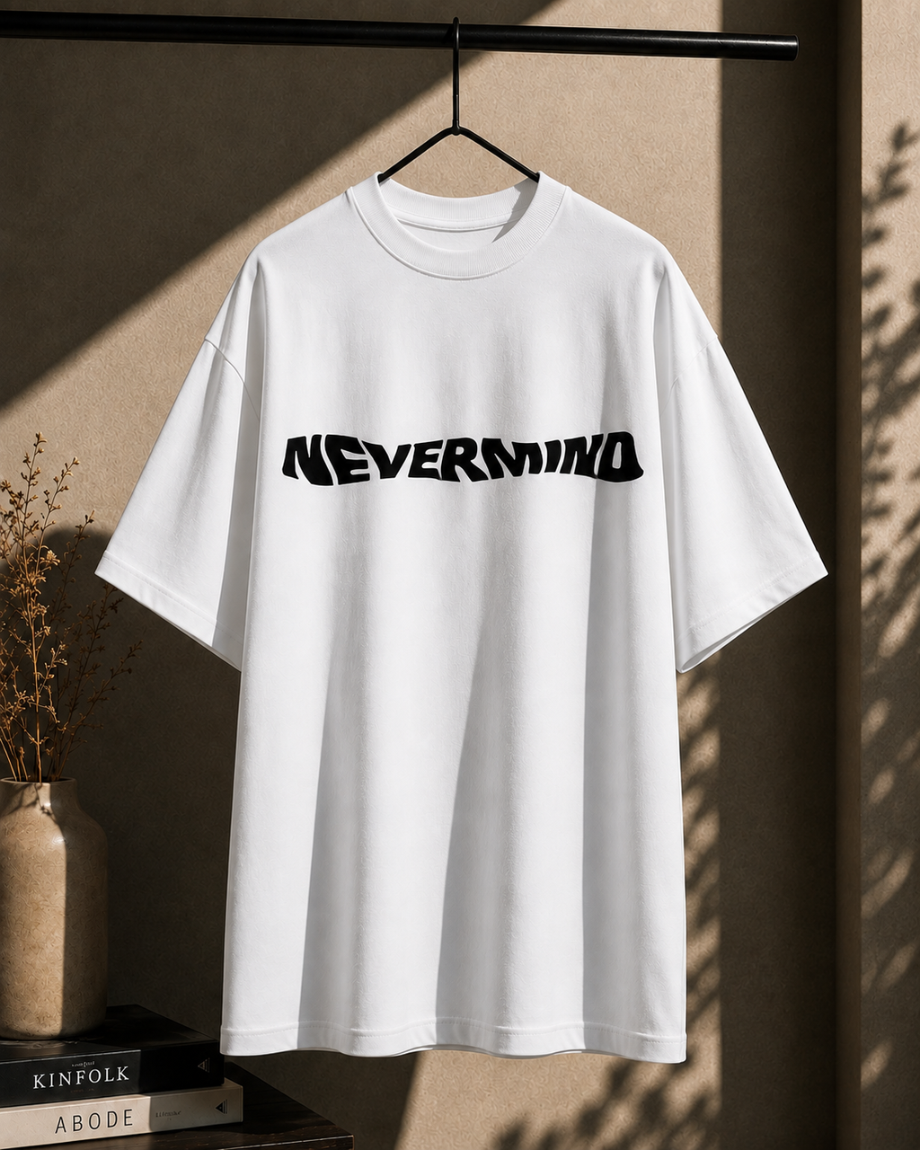 A white oversized short-sleeve T-shirt hanging on a black hanger against a textured beige wall, with a bold black 'NEVERMIND' graphic across the chest. Decorative books, a vase with dried stems, and leaf shadows are visible around it.