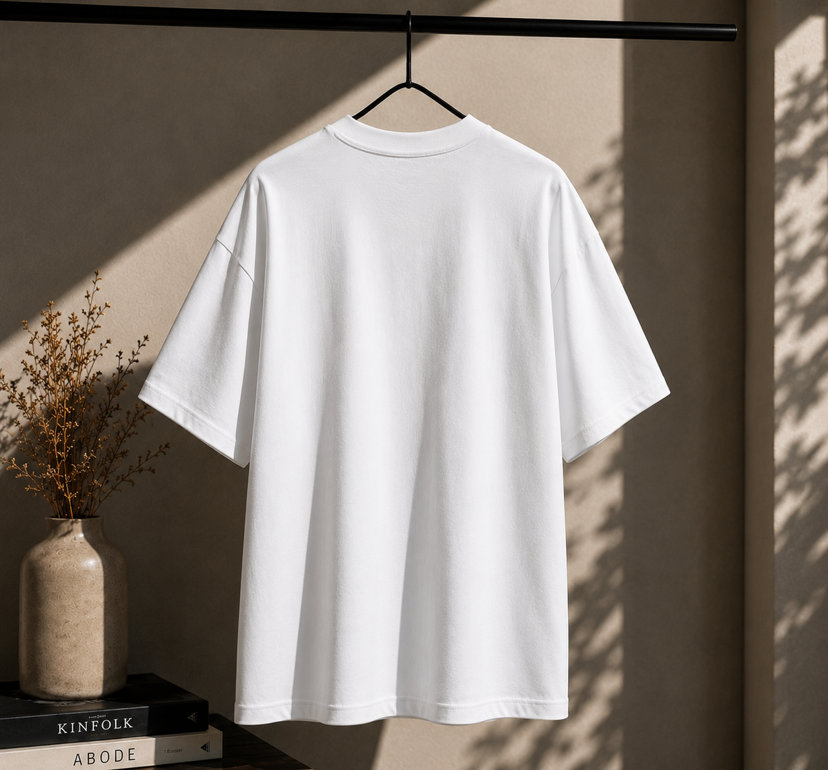 A plain white short-sleeve T-shirt hanging on a black hanger against a warm beige wall, with soft sunlight casting shadows and dried flowers in a vase nearby.