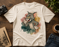 A cream-colored graphic t-shirt featuring Tom and Jerry's character Tom in a watercolor splash design, styled in a flat-lay on a dark wood table with a camera, notebook, coffee cup, and jeans.