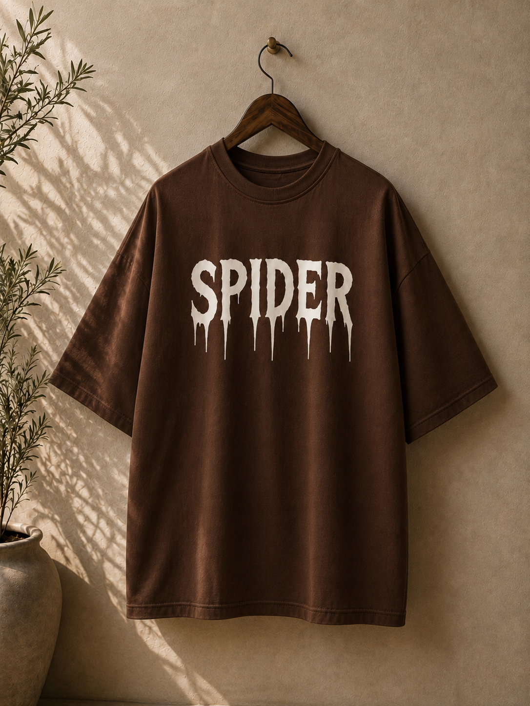 A brown oversized short-sleeve T-shirt hanging on a wooden hanger against a textured beige wall, with a large white dripping 'SPIDER' graphic across the chest and a potted plant casting shadows on the left.