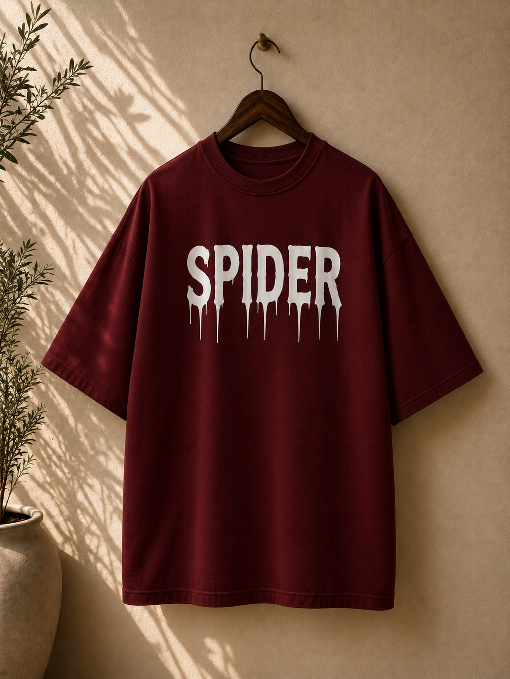 A burgundy short-sleeve t-shirt hanging on a wooden hanger against a beige wall, featuring the word 'SPIDER' printed in a dripping white font across the chest.