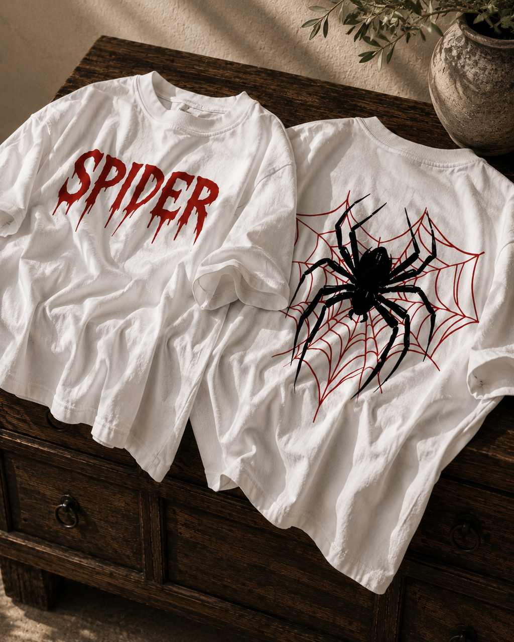 Two white graphic T-shirts displayed on a dark wooden chest. The front shirt features the word 'SPIDER' in a dripping red font, and the back shirt features a large black spider graphic over a red spiderweb.