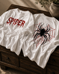 Two white graphic T-shirts displayed on a dark wooden chest. The front shirt features the word 'SPIDER' in a dripping red font, and the back shirt features a large black spider graphic over a red spiderweb.