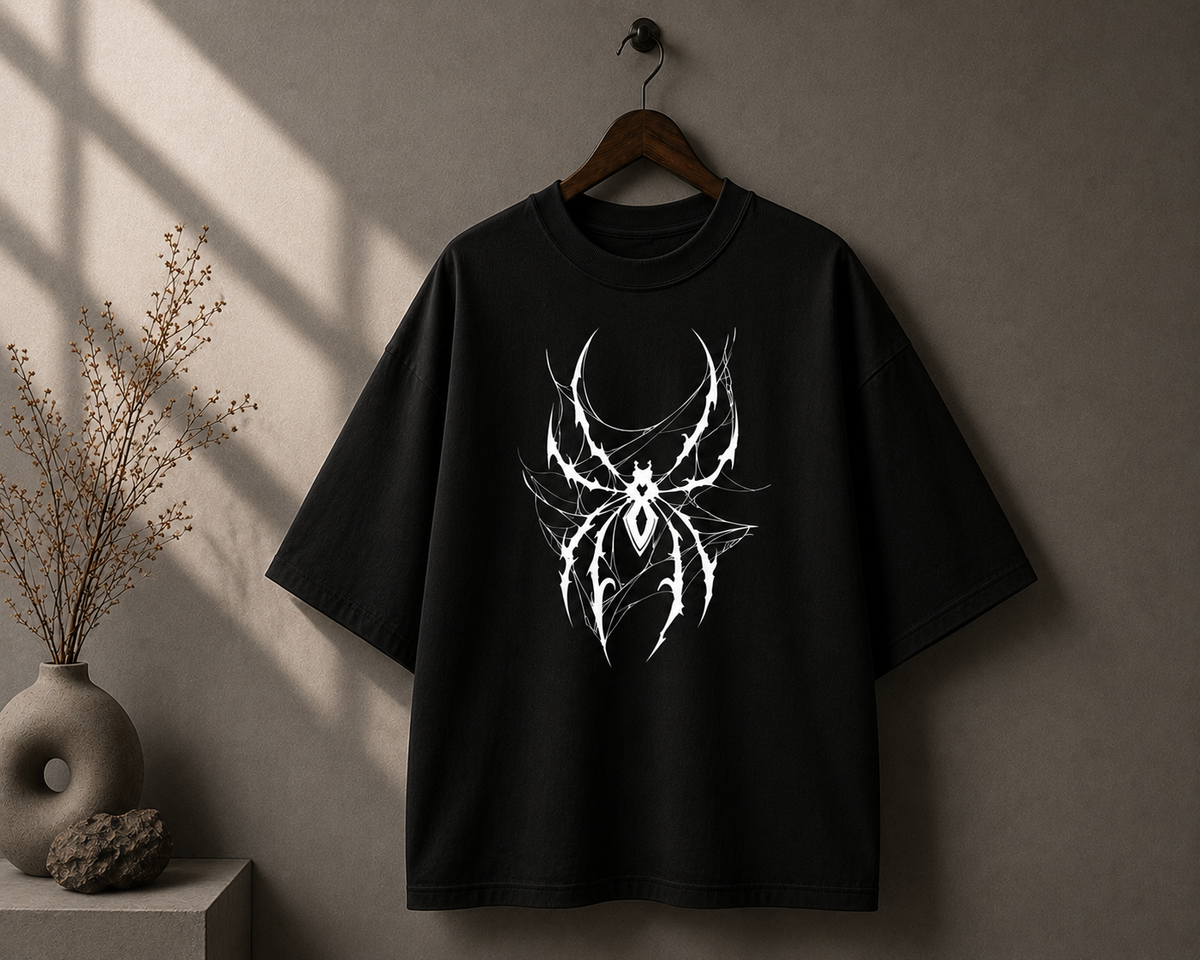 A black oversized short-sleeve T-shirt hanging on a wooden hanger against a textured gray wall, featuring a large white stylized spider graphic on the chest. A vase with dried branches sits on a side table to the left.