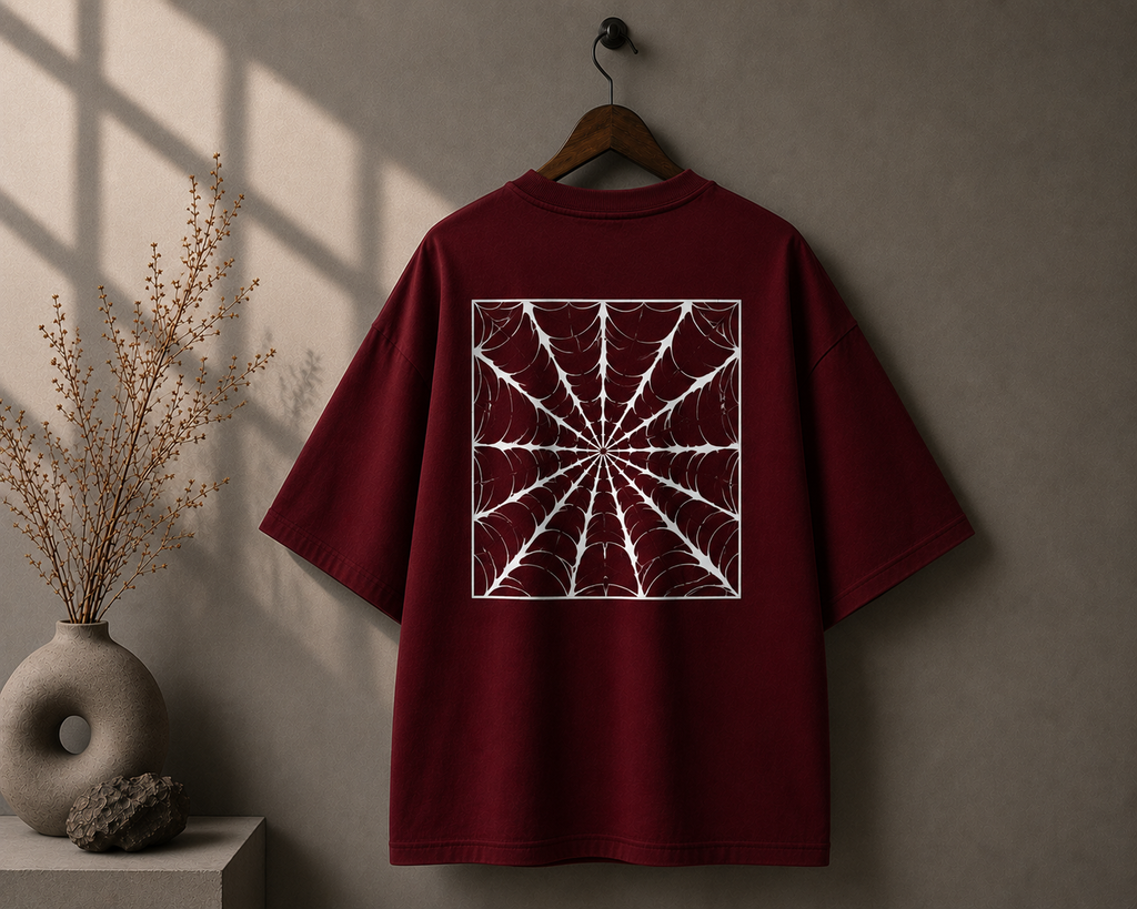 A burgundy short-sleeve T-shirt hanging on a wooden hanger against a textured gray wall, featuring a large white spiderweb square graphic printed on the back. The scene is styled with a ceramic vase of dried branches on the left and soft window shadows cast across the wall.